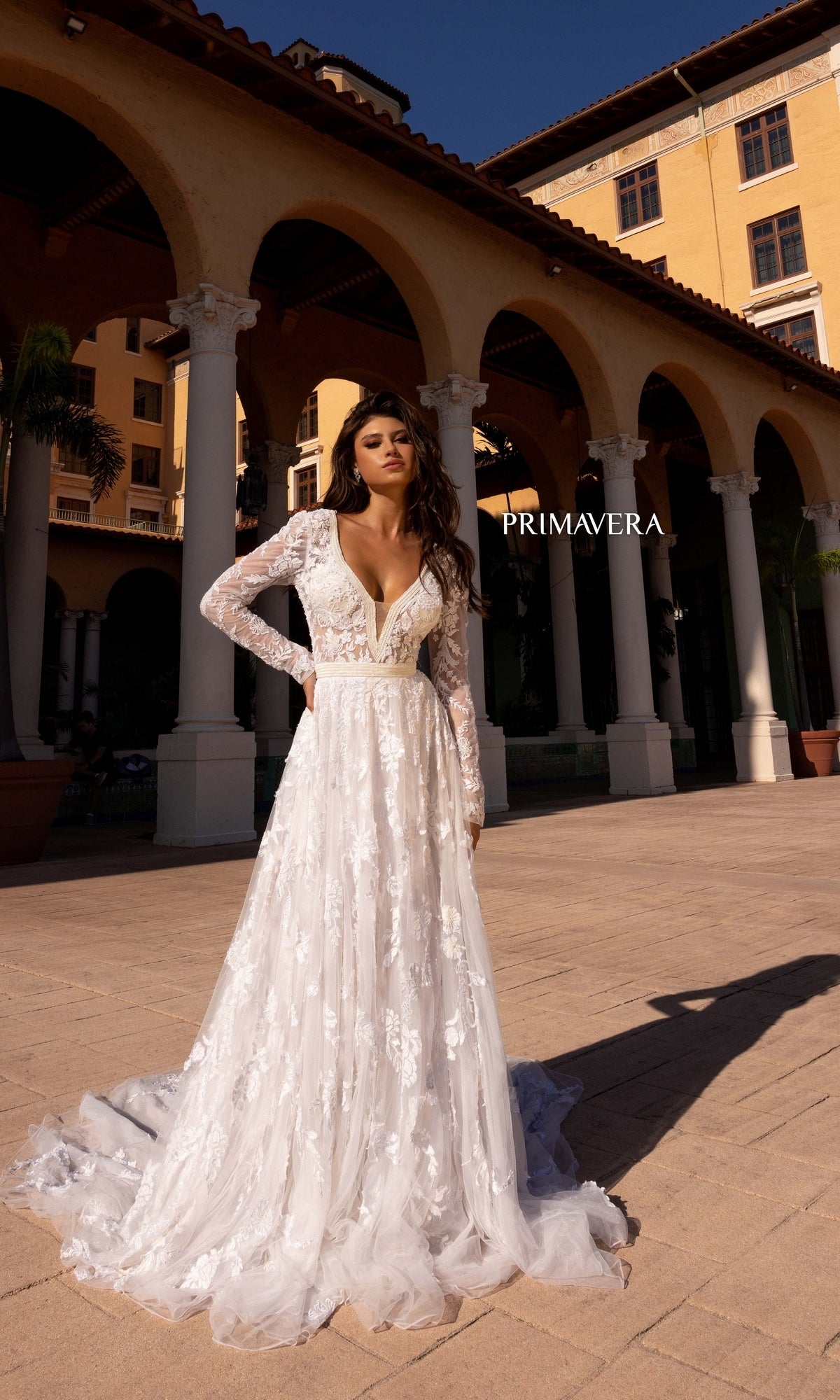 A woman wearing the Long Wedding Dress 11111 by Primavera stands outdoors in front of a Mediterranean-style building, with